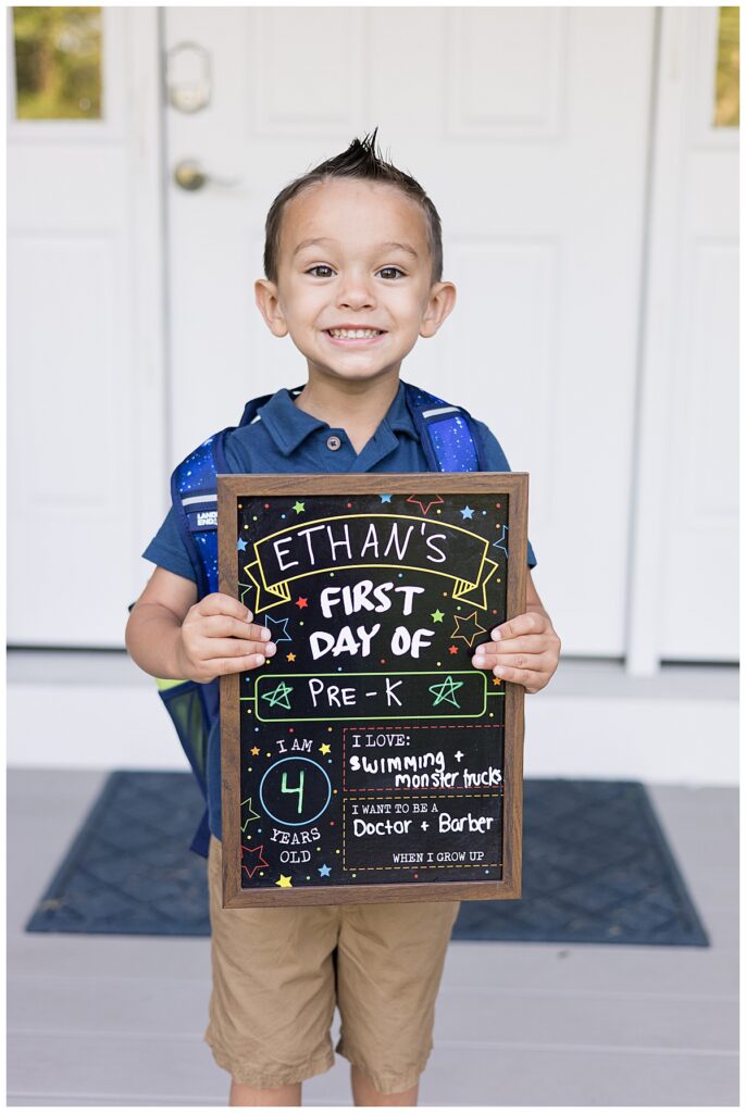 A close-up of Ethan standing in front of a white door, smiling widely and holding the chalkboard sign. Photo taken in Westport, MA by family photographer Lorie-Lyn Photography. Watch Me Grow Collection