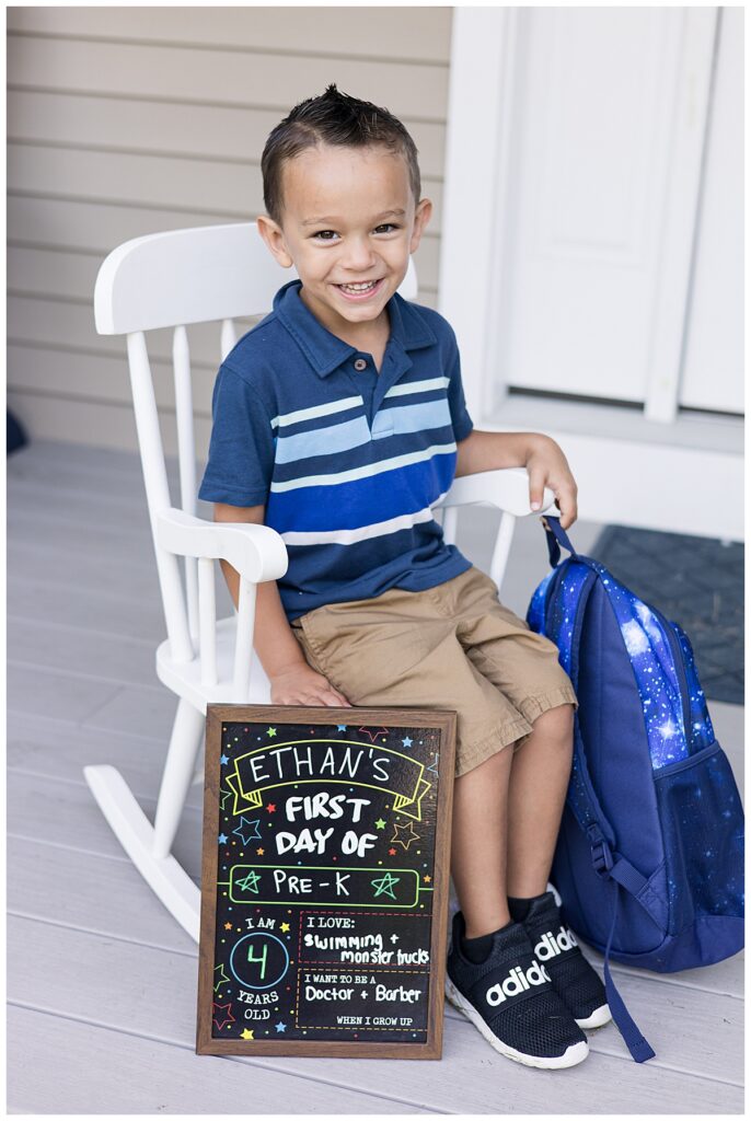 A close-up of Ethan sitting in front of a white door, smiling widely and holding the chalkboard sign. Photo taken in Westport, MA by family photographer Lorie-Lyn Photography. Watch me Grow Collection
