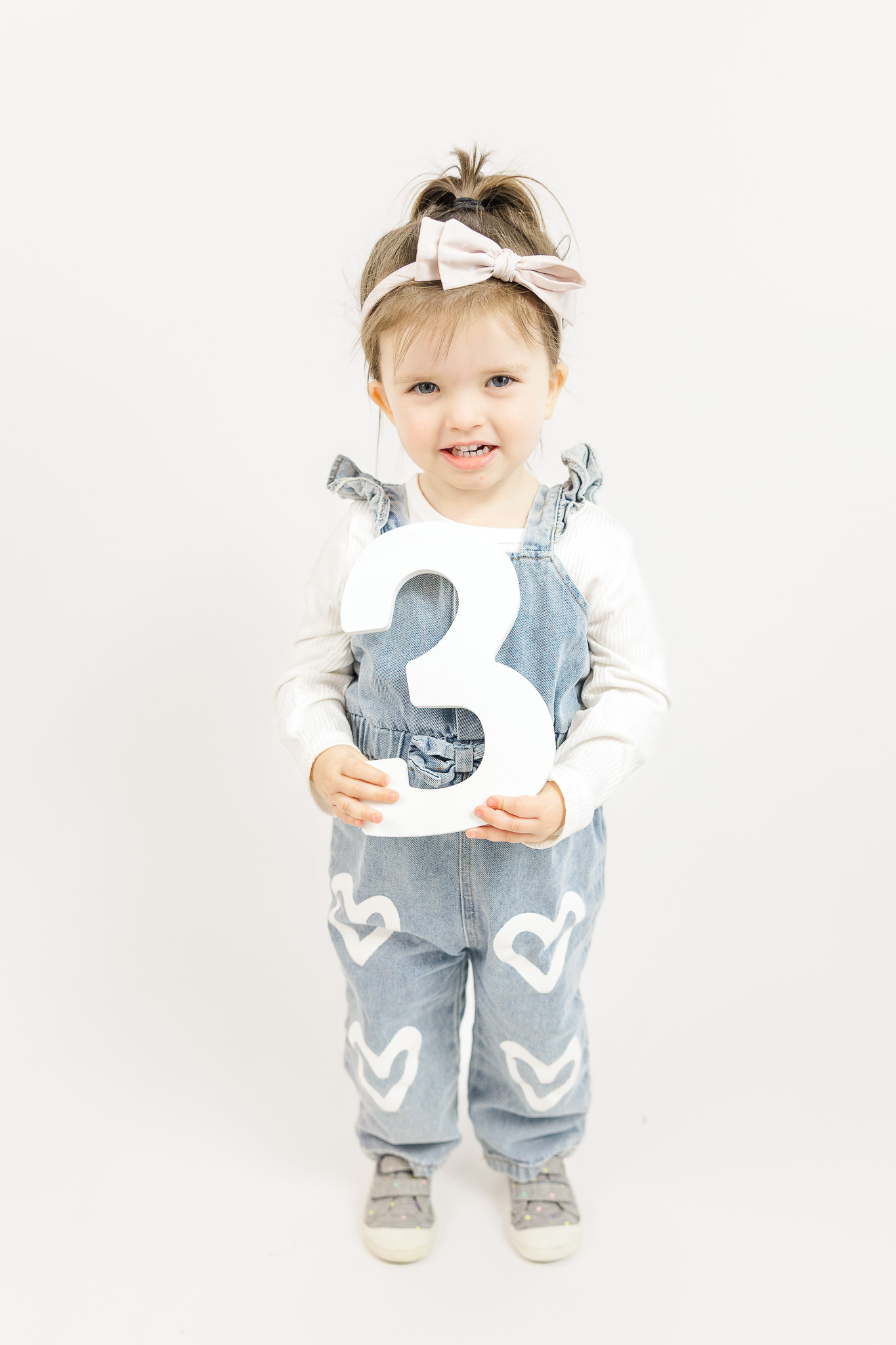 Smiling 3-year-old girl in denim overalls and a bow headband standing against a white background