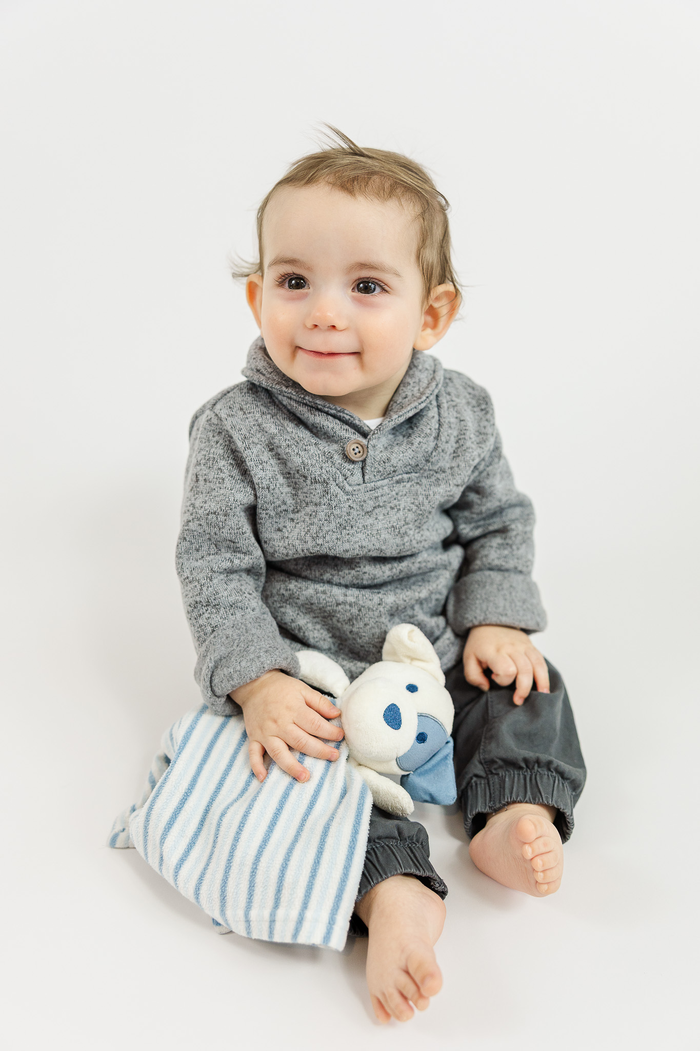 Smiling 8-month-old baby boy sitting on a soft blanket holding a plush bunny, photographed in natural light.